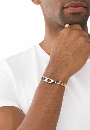 Goldene Kettenarmband mit silbernem Beschlägen und einem auffälligen Schnallendesign. Am Handgelenk getragen, hebt es seine Ästhetik aus gemischten Metallen hervor.