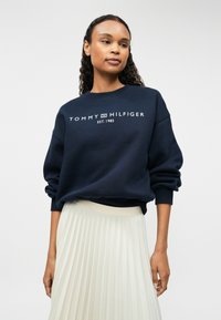 Sweatshirt bleu marin cu guler rotund, având un logo alb care spune "TOMMY HILFIGER EST. 1985" pe partea din față. Material moale, texturat.