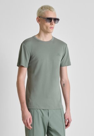 Uomo con capelli biondi corti che indossa occhiali da sole scuri, t-shirt verde chiaro, pantaloni abbinati e mostra un piccolo tatuaggio sull'avambraccio sinistro.
