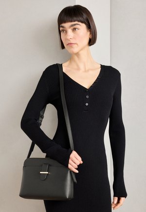 Žena s krátkými tmavými vlasy v černých žebrovaných šatech držící černou koženou kabelku přes rameno na neutrálním pozadí.