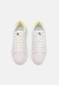 Pair of white low-top sneakers with white laces, yellow heel tabs, and black and yellow "CK" Calvin Klein logos on the tongues.