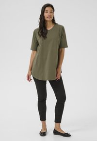 Camiseta de corte oversized en verde oliva con mangas cortas y dobladillo redondeado, combinada con pantalones ajustados negros. Unos zapatos planos negros completan el conjunto.
