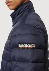 Giacca imbottita blu navy con colletto alto, caratterizzata da sezioni trapuntate e una patch nera con testo bianco e rosso sulla spalla sinistra.