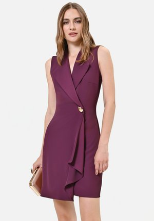 Femme portant une robe sans manches violet foncé avec un détail asymétrique de boutons, tenant une pochette rectangulaire métallique, sur fond uni.