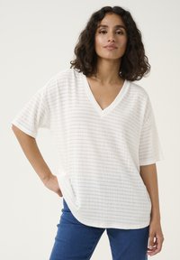 White short-sleeve V-neck top with textured fabric, featuring a side pocket and a rounded hemline. Styled with blue jeans.