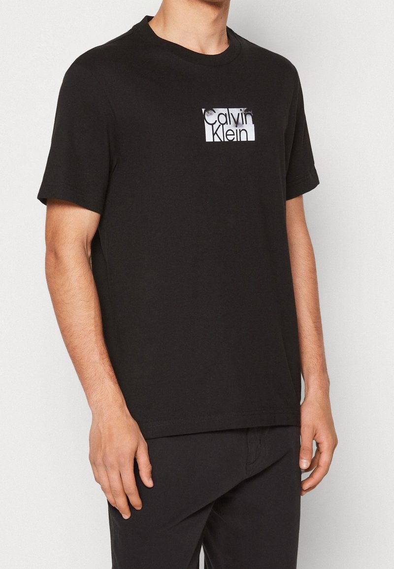 Camiseta negra de algodón con mangas cortas, con un logo blanco de Calvin Klein en un pequeño parche rectangular en el pecho.