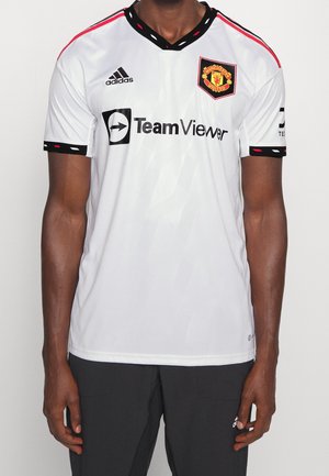 Man draagt een wit Manchester United voetbalshirt met zwarte en rode details, Adidas- en TeamViewer-logo's, gecombineerd met een zwarte broek.