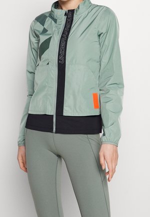 Veste de sport vert clair avec une fermeture éclair sur toute la longueur à l'avant, logo noir et poche latérale. Présente des poignets élastiques et un accent orange contrastant.