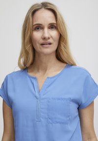 Světle modrá blůzka s krátkým rukávem, V-výstřihem a kapsou na prsou. Má hladkou tkaninu a jemný šití podél výstřihu.