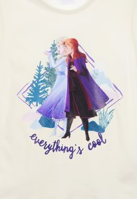 Top de tirantes crema que presenta dos personajes ilustrados contra un fondo de árboles y montañas. El texto debajo dice "todo está bien" en color morado.