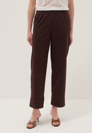 Femme portant un pantalon droit marron foncé à taille élastique associé à des sandales à talons ouvertes de couleur beige sur un fond uni.
