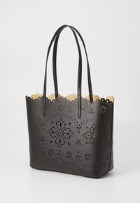 Black perforated tote bag with intricate floral cutouts, smooth texture, and scalloped edge; features dual handles and a beige interior.