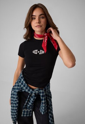 Ung kvinde iført en sort Guess Jeans t-shirt, rød halstørklæde og en blå ternet skjorte bundet om taljen, poserer mod en grå baggrund.