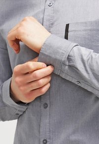 Camisa gris abotonada en una tela lisa; cuenta con un bolsillo en el pecho con un detalle oscuro, puños ajustables y botones grises.