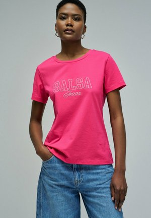 Mujer con una camiseta rosa brillante de "SALSA Jeans" y pantalones vaqueros azules, de pie con una mano en el bolsillo contra un fondo claro y liso.