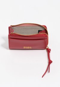 Czerwony skórzany portfel z złotym zamkiem, z teksturowanym wnętrzem i dekoracyjnym frędzlem. Logo wytłoczone z przodu.