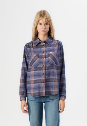Femme portant une chemise à carreaux bleu et violet avec des poches poitrine et un jean en denim bleu clair, se tenant contre un fond uni.