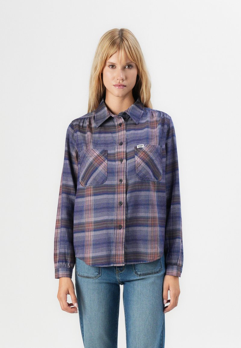 Woman wearing a blue and purple plaid button-up shirt with chest pockets and light blue denim jeans, standing against a plain background.
