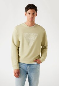 Lichtgroen sweatshirt met ribgebreide manchet en halslijn. Voorzien van een verhoogd wit logo op een driehoekig ontwerp. Gecombineerd met lichtblauwe spijkerbroek.