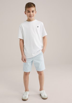 Witte T-shirt met korte mouwen met een zwart logo, gecombineerd met lichtblauwe shorts. Het schoeisel bestaat uit wit-grijze sneakers met textuurzolen.