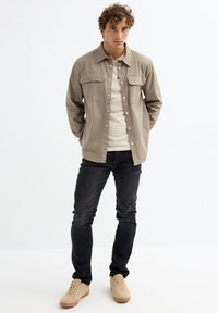 Camisa de botón beige con dos bolsillos en el pecho sobre una camiseta clara, combinada con jeans negros y zapatillas beige. Ajuste casual, textura de tela suave.