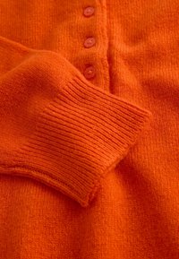 Oranger Strickpullover mit gerippten Bündchen und fünf vorderen Knöpfen in passendem Orangeton. Weiche Textur mit mittlerem Gewicht.