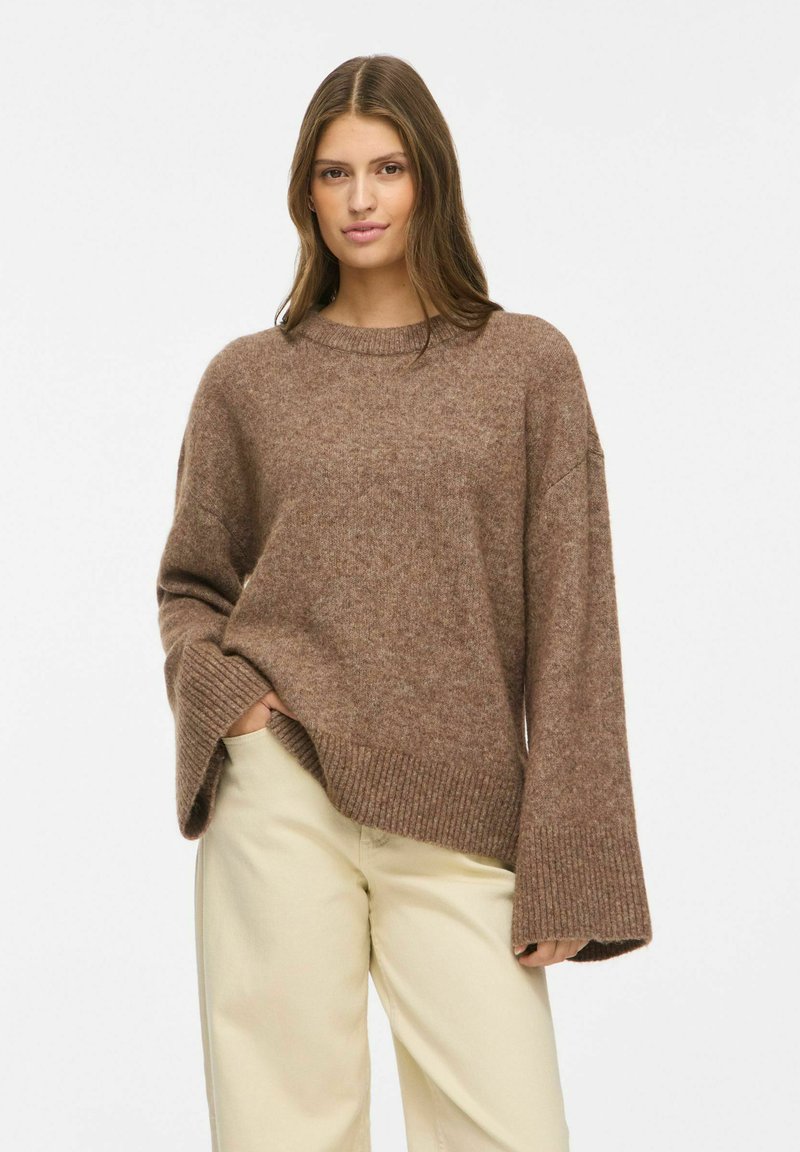 Jeune femme debout, une main dans la poche, portant un pull marron ample à manches larges et un pantalon crème sur un fond uni.