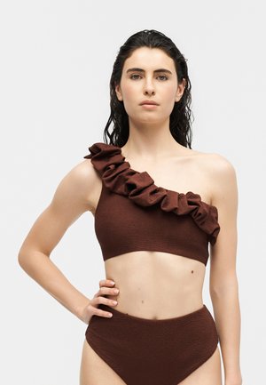 Femme portant un bikini asymétrique marron foncé avec une bretelle à volants et un bas taille haute, debout la main sur la hanche devant un fond uni.