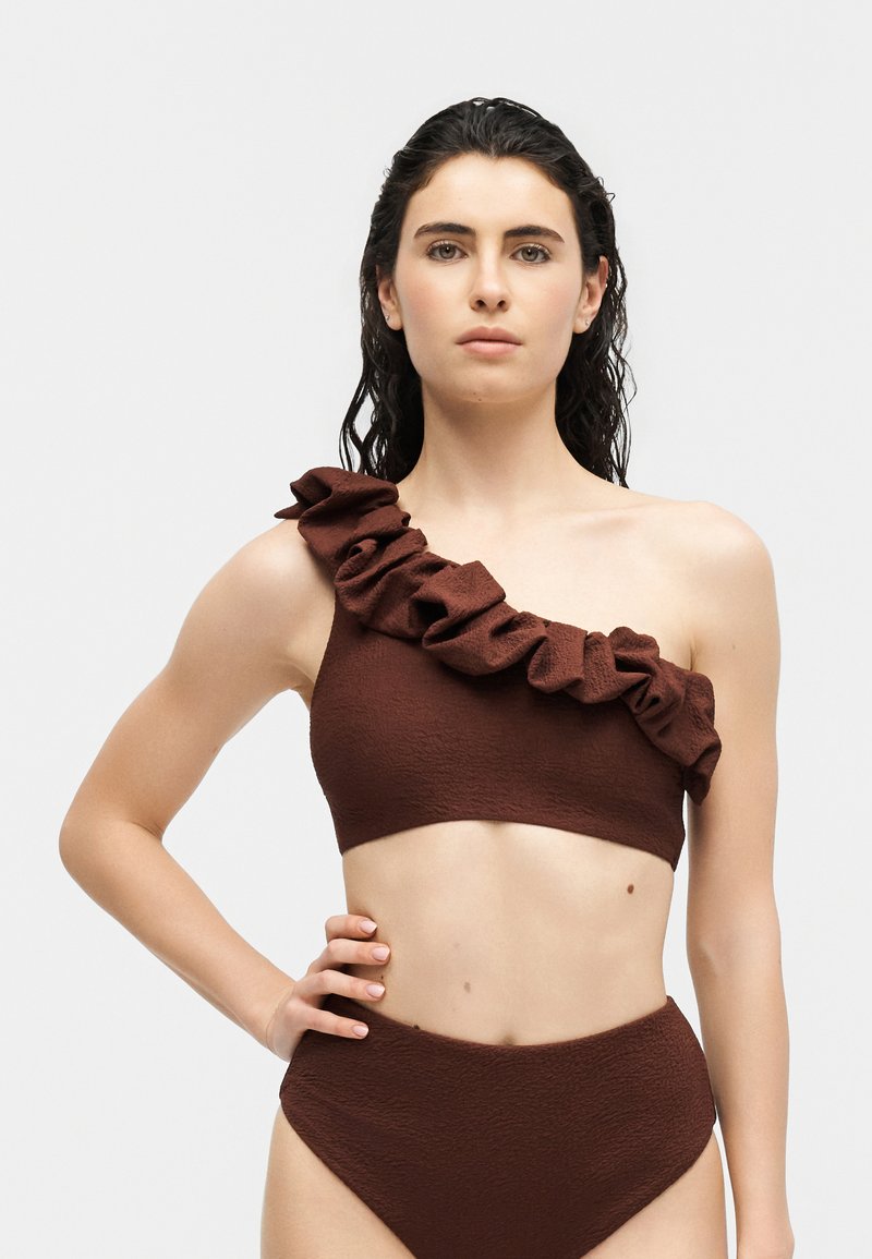 Γυναίκα που φοράει μαγιό μπικίνι σκούρου καφέ χρώματος με έναν ώμο, με βολάν στο λουρί και σλιπ με ψηλή μέση, στέκεται με το χέρι στη μέση μπροστά σε απλό φόντο.