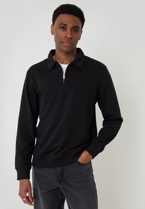 Homme aux cheveux courts et bouclés portant un pull noir à fermeture éclair quart-zip et un jean gris foncé, debout avec une main dans la poche.