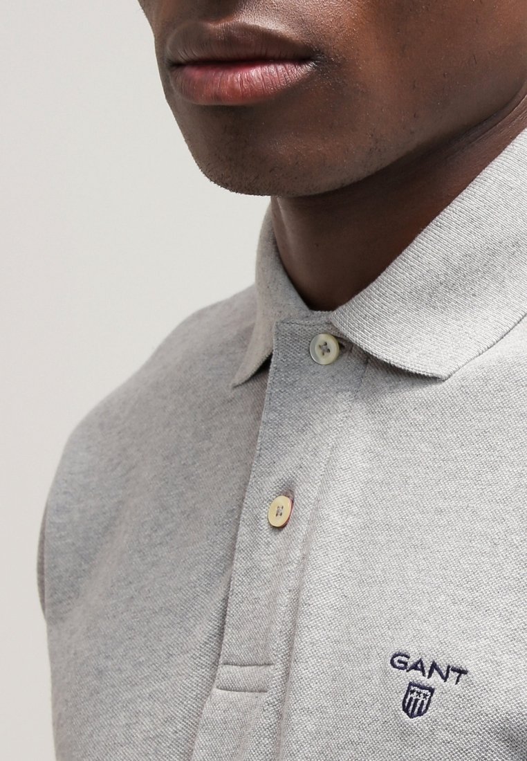 Gros plan d'une personne portant un polo GANT gris clair avec deux boutons fermés et un logo brodé sur la poitrine.