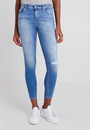 Vrouw draagt lichtblauwe beschadigde skinny jeans en witte sneakers, staand tegen een effen grijze achtergrond.