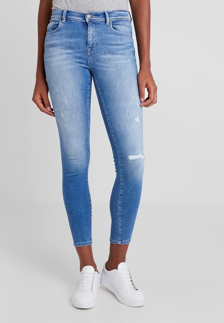 Femme portant un jean skinny bleu clair déchiré et des baskets blanches, debout devant un fond gris uni.