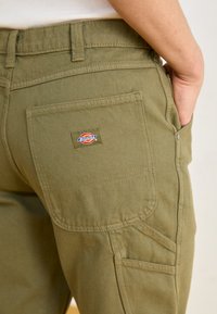 Olive green cargo pants made of cotton with a textured finish, featuring a prominent back pocket and a visible logo patch.