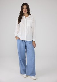 Weißes, langärmliges Hemd mit Knopfleiste und Brusttasche, kombiniert mit hellblauen, weit geschnittenen Jeans und weißen Turnschuhen. Glatte Stofftextur.