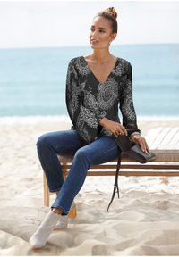 Schwarz-weiß gemusterte Langarmbluse mit V-Ausschnitt, kombiniert mit blauen Jeans und hellgrauen Knöchelstiefeln mit Holzabsatz; hält eine kleine schwarze Clutch.