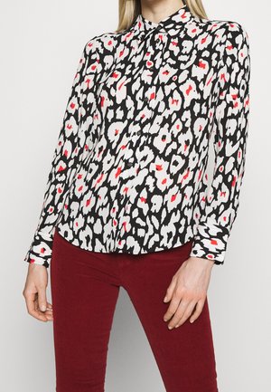 Femme portant un chemisier à manches longues à motifs noir et blanc avec des accents rouges et un pantalon rouge foncé ajusté, debout devant un fond uni.