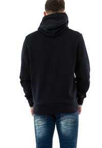 Granatowa bluza z kapturem z dużym kapturem, miękkim materiałem, ściągaczami na rękawach i u dołu. Noszona z niebieskimi dżinsami, z widocznymi przetarciami z tyłu.
