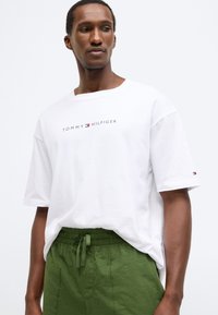 Λευκό βαμβακερό μπλουζάκι με στρογγυλή λαιμόκοψη και κοντά μανίκια, με εκτυπωμένο λογότυπο "TOMMY HILFIGER" σε μαύρο και κόκκινο. Συνδυάζεται με πράσινο παντελόνι.
