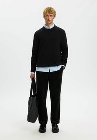 Noor mees mustas sviitrisse, helesinises särgis, mustades pükstes ja kingades hoiab tumedat halli kottide kotti ühe lihtsa valge tausta ees.