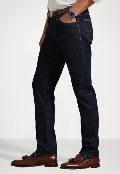 Sötét farmernadrág, slim fit szabással, kontrasztos sárga varrással, öt zsebes kialakítással, barna papucscipővel és órával párosítva.