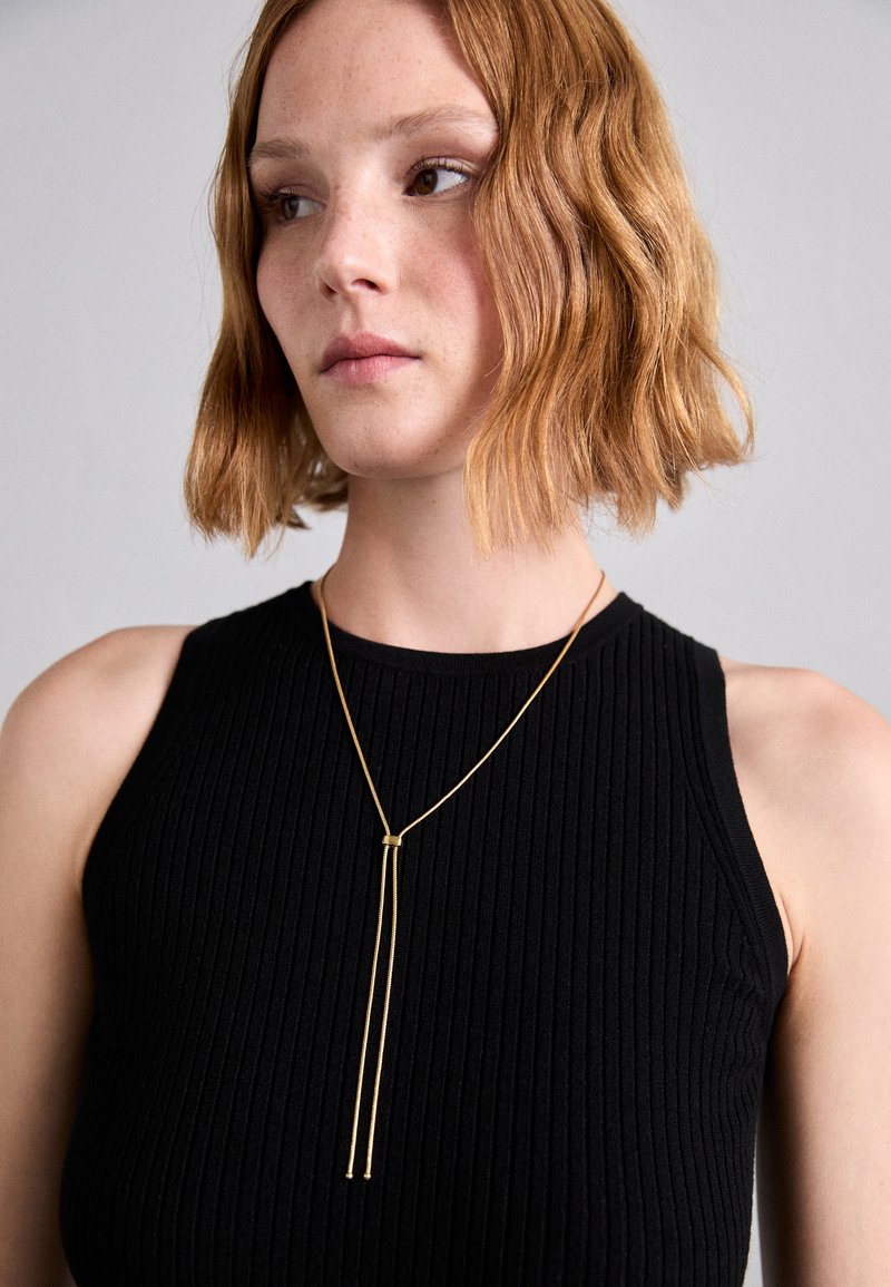 Kultaketjun kaulakoru, jossa on pitkä ketju, pystysuuntainen muotoilu ja kaksi roikkuvaa narua. Vaatteena musta ribbitoppi.