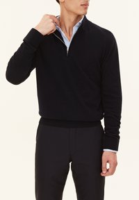 Pull marine en matériau doux avec un col montant et une fermeture éclair à l'avant. Présente des poignets et un ourlet ajustés, porté par-dessus une chemise à col bleue claire.