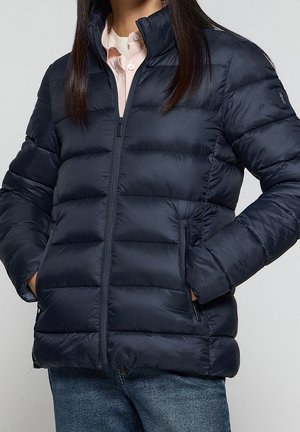 Femme portant une doudoune matelassée bleu marine avec fermeture éclair devant et les mains dans les poches, associée à un jean bleu clair et une chemise rose clair.