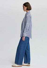 Camisa de rayas en azul y blanco, combinada con jeans anchos azul y zapatillas blancas. La camisa tiene mangas oversized y un corte relajado.
