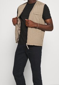Chaleco acolchado beige con cierre frontal, bolsillo en el lado izquierdo del pecho y fabricado con un material ligero. Se lleva sobre una camiseta negra y pantalones oscuros.