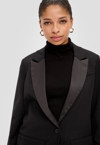 Black blazer with satin lapels, structured design, single button closure, two front pockets, worn over a black turtleneck.