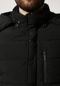 Czarna kurtka puchowa z wysokim kołnierzem, zatrzaskami, kieszenią na zamek i teksturowaną tkaniną o delikatnym połysku.