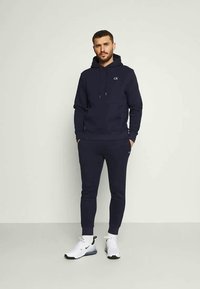 Hanorac bleumarin cu șnururi și buzunar frontal, asortat cu pantaloni jogger. Are un mic logo "ck". Este purtat cu adidași albi.