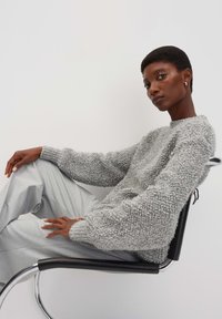 Maglione grigio chiaro a maglia strutturata con scollo rotondo e maniche lunghe e voluminose. La modella posa seduta su una sedia nera elegante, indossando pantaloni grigi.